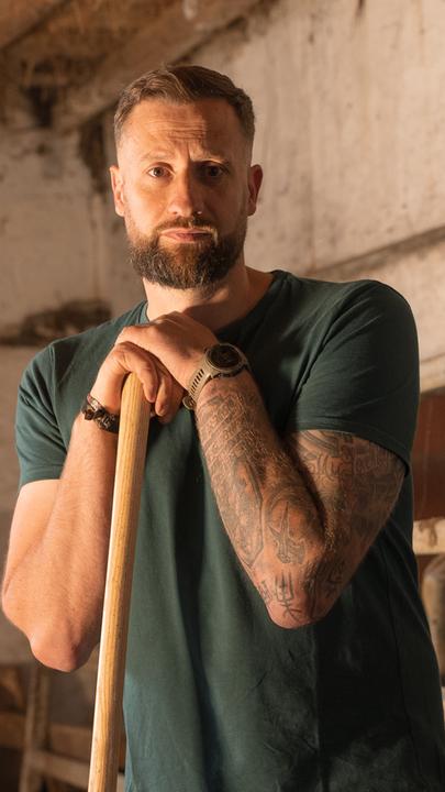 Christoph Rothhaupt, ein Mann mit braunen Haaren und Bart, steht in einem Stall. Er stützt auf einer Mistgabel und schaut mit traurigem Blick in die Kamera. Sein linker Arm ist tätowiert, er trägt eine Uhr. Im Hintergrund ist eine verschmutzte Wand und eine Decke mit Holzbalken, zwischen denen Stroh raus ragt.