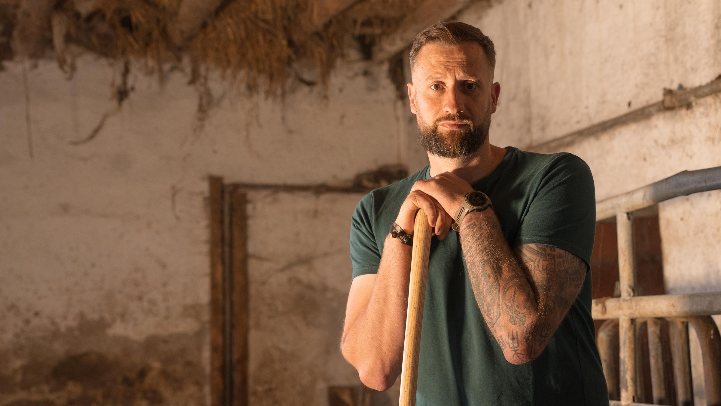 Christoph Rothhaupt, ein Mann mit braunen Haaren und Bart, steht in einem Stall. Er stützt auf einer Mistgabel und schaut mit traurigem Blick in die Kamera. Sein linker Arm ist tätowiert, er trägt eine Uhr. Im Hintergrund ist eine verschmutzte Wand und eine Decke mit Holzbalken, zwischen denen Stroh raus ragt.