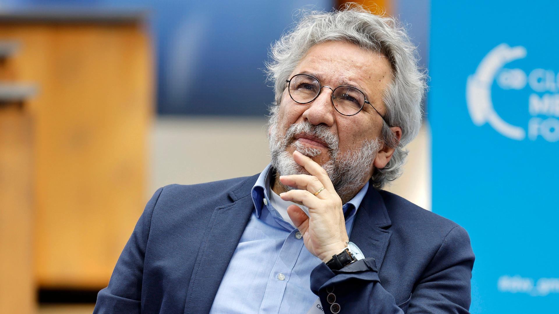 Foto eines Mannes mit weißen Locken, graumeliertem Bart und runder Metall-Brille, der sich nachdenklich mit der Hand ans Kinn fasst.