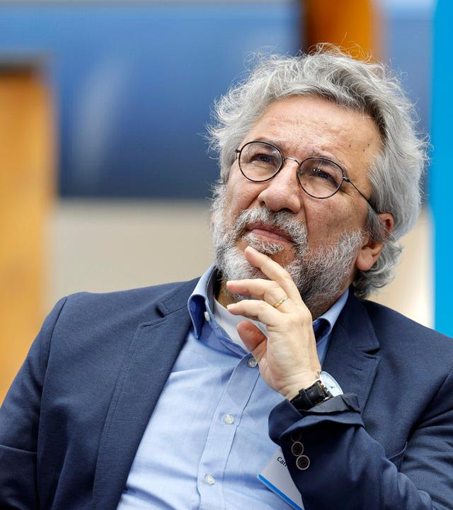 Foto eines Mannes mit weißen Locken, graumeliertem Bart und runder Metall-Brille, der sich nachdenklich mit der Hand ans Kinn fasst.