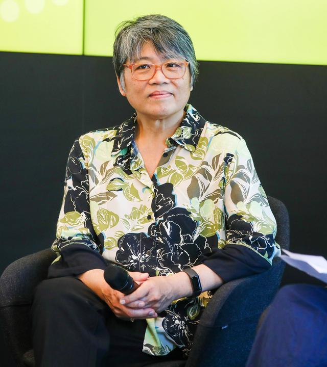 Foto einer asiatisch aussehenden Frau mittleren Alters mit kurzen graumelierten Haaren und Brille, die ein grün, weiß, grau gemustertes Hemd trägt.