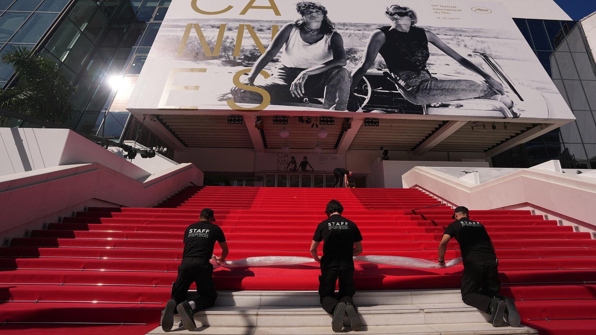 Drei schwarz gekleidete Männer rollen auf den Treppen zum Kino in Cannes einen roten Teppich ab.