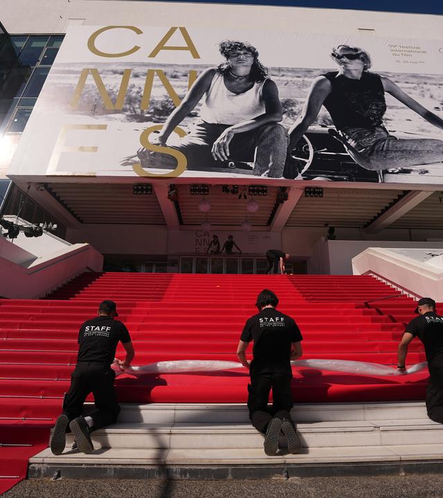 Drei schwarz gekleidete Männer rollen auf den Treppen zum Kino in Cannes einen roten Teppich ab.