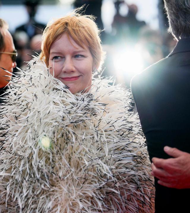 Eine Frau mit kurzen rötlichen Haaren und bekleidet mit einer schicken aufgepufften weiß-schwarzen Zotteljacke steht zwischen zwei Männdern in Anzügen, die nur von hinten zu sehen sind, und dreht sich zum Betrachter um.