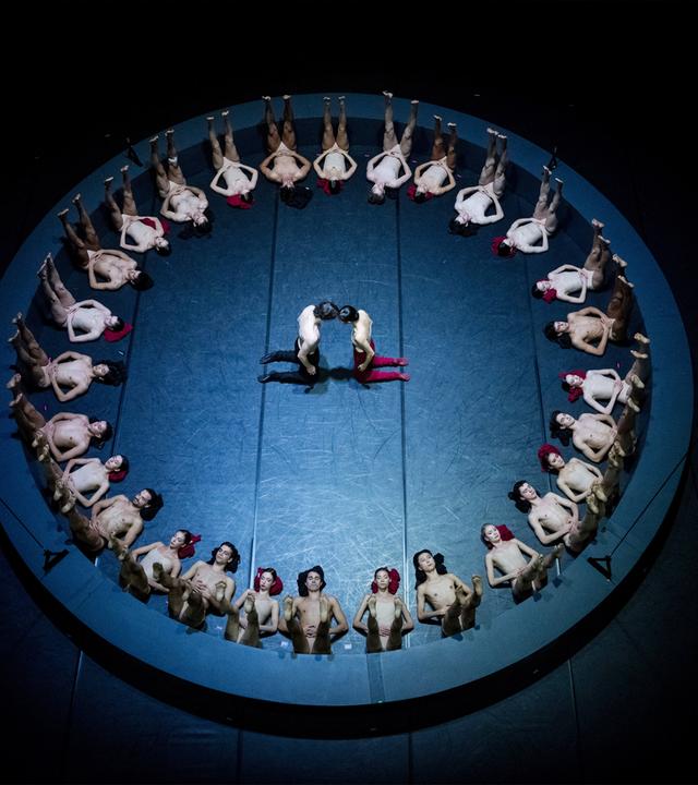 Menschen liegen in einem Kreis auf dem Boden. In der Mitte des Kreises knien sich zwei Menschen gegenüber und berühren sich mit den Köpfen.