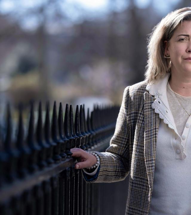 Porträtfoto einer Frau mit schulterlangen Haaren, die einen karierten Anzug trägt. Sie steht an einem schmiedeeisernen Zaun und blickt ernst in die Kamera.