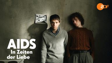 Heiko (Evangelos Tsarkowistas) und Dirk (Benjamin Viziotis) stehen nebeneinander vor einer grauen Wand mit einer Schwarz-Weiß-Zeichnung. 