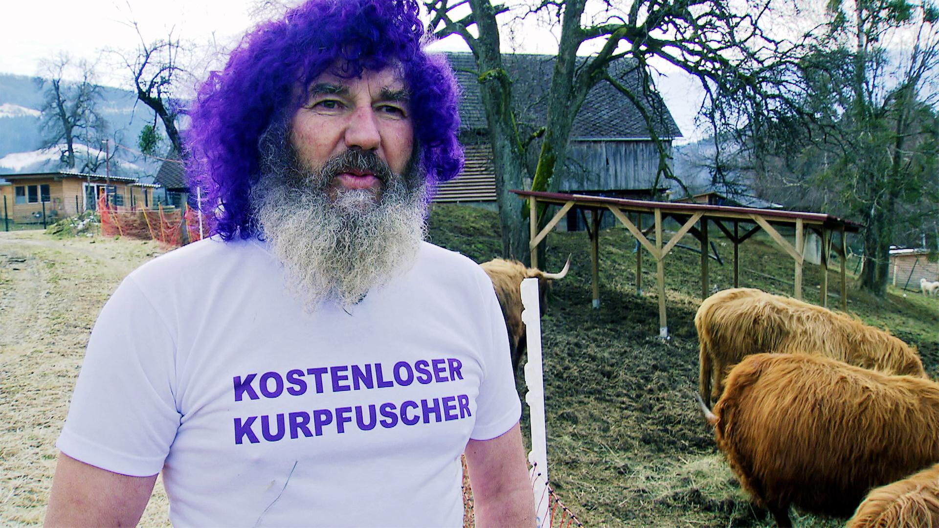 Das Bild zeigt einen Mann mit einem markanten Aussehen. Er hat einen dichten, grauen Bart und trägt eine hellgraue T-Shirt mit dem Aufdruck „KOSTENLOSER KURPFUSCHER“ in lila Buchstaben. Seine Haare sind auffällig lila gefärbt und lockig. Der Mann steht in einem Außenbereich, hinter ihm sind mehrere Hochlandrinder zu sehen, die dichtes, hellbraunes Fell haben. Die Umgebung ist ländlich, mit einem leicht bewaldeten Hintergrund und einem Holzgebäude, das in der Ferne sichtbar ist. Um den Bereich sind Absperrungen zu erkennen, und der Boden ist überwiegend braun mit kurzer Vegetation. Der Himmel ist bewölkt, was auf schlechtes Wetter hindeutet. Die gesamte Szene vermittelt eine entspannte, jedoch ungewöhnliche Atmosphäre, die mit der Thematik der Energetik in Verbindung steht, die im Text erwähnt wird.