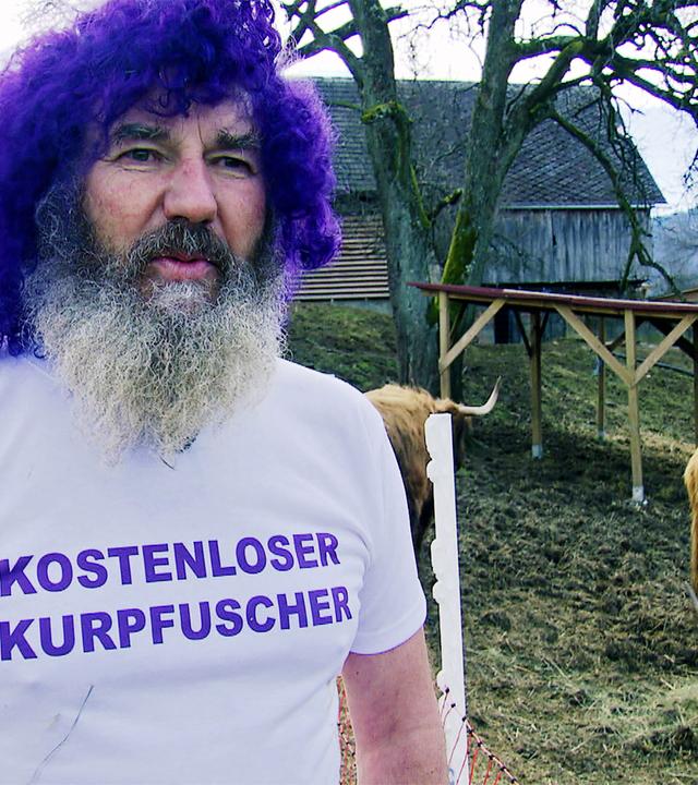 Das Bild zeigt einen Mann mit einem markanten Aussehen. Er hat einen dichten, grauen Bart und trägt eine hellgraue T-Shirt mit dem Aufdruck „KOSTENLOSER KURPFUSCHER“ in lila Buchstaben. Seine Haare sind auffällig lila gefärbt und lockig. Der Mann steht in einem Außenbereich, hinter ihm sind mehrere Hochlandrinder zu sehen, die dichtes, hellbraunes Fell haben. Die Umgebung ist ländlich, mit einem leicht bewaldeten Hintergrund und einem Holzgebäude, das in der Ferne sichtbar ist. Um den Bereich sind Absperrungen zu erkennen, und der Boden ist überwiegend braun mit kurzer Vegetation. Der Himmel ist bewölkt, was auf schlechtes Wetter hindeutet. Die gesamte Szene vermittelt eine entspannte, jedoch ungewöhnliche Atmosphäre, die mit der Thematik der Energetik in Verbindung steht, die im Text erwähnt wird.