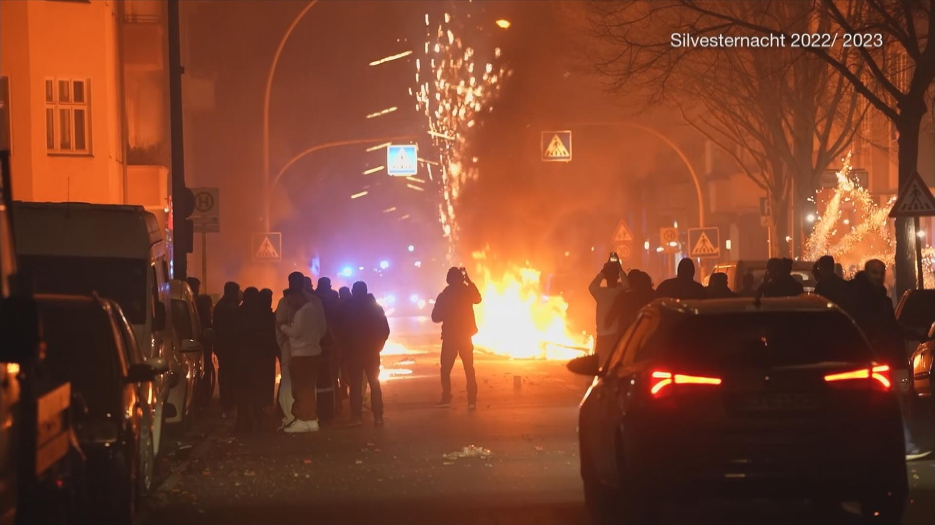 Flammen und Böller auf einer Straße. Mehrere Menschen stehen mit dem Handy filmend davor.
