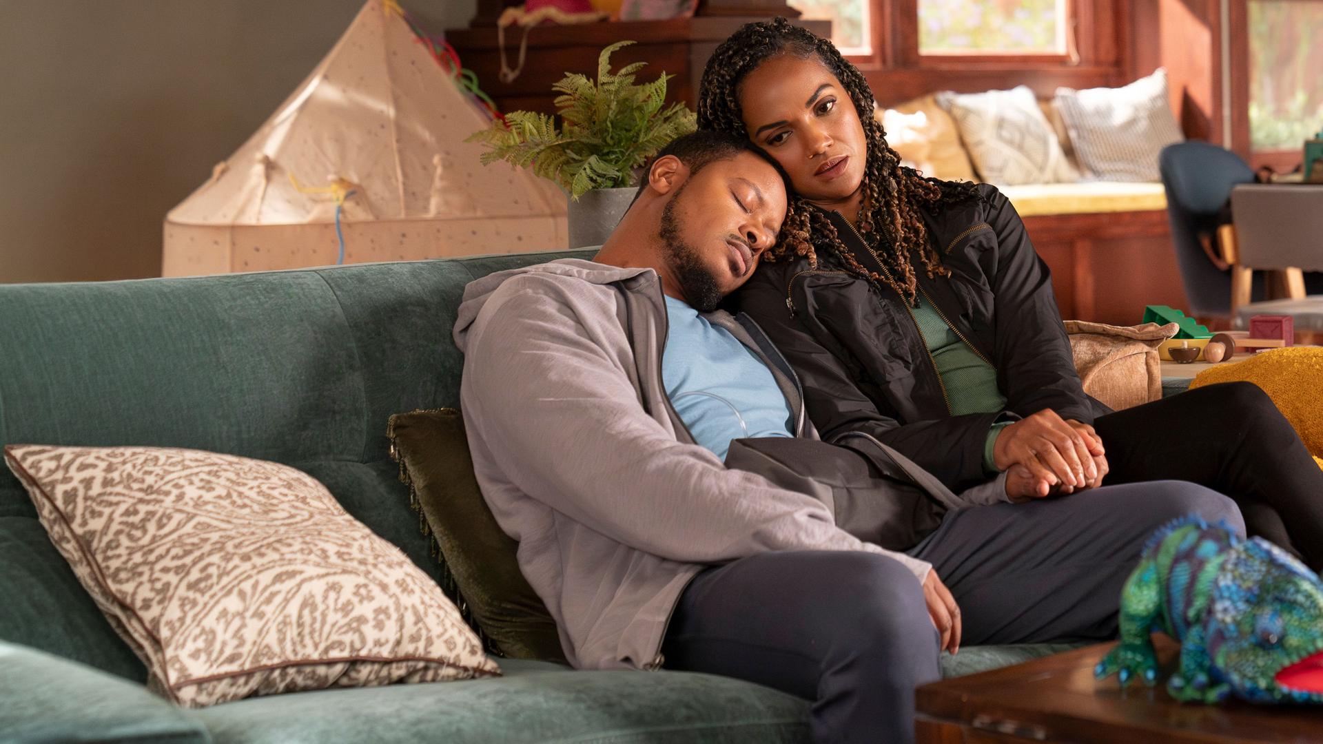 "The Rookie - Chaos": James Murray (Arjay Smith) und Detective Harper (Mekia Cox) sitzen zusammen auf einem Sofa in einem Wohnzimmer. Beide tragen Alltagskleidung. James hat seinen Kopf auf Harpers Schulter gelegt und sie schmiegt wiederum ihren Kopf an seinen an. Seine Augen sind geschlossen, während sie nach vorn blickt.