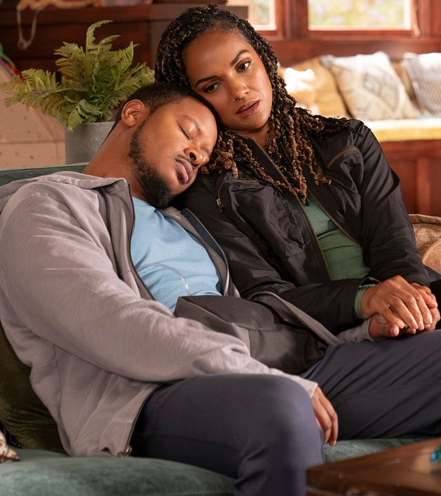 "The Rookie - Chaos": James Murray (Arjay Smith) und Detective Harper (Mekia Cox) sitzen zusammen auf einem Sofa in einem Wohnzimmer. Beide tragen Alltagskleidung. James hat seinen Kopf auf Harpers Schulter gelegt und sie schmiegt wiederum ihren Kopf an seinen an. Seine Augen sind geschlossen, während sie nach vorn blickt.