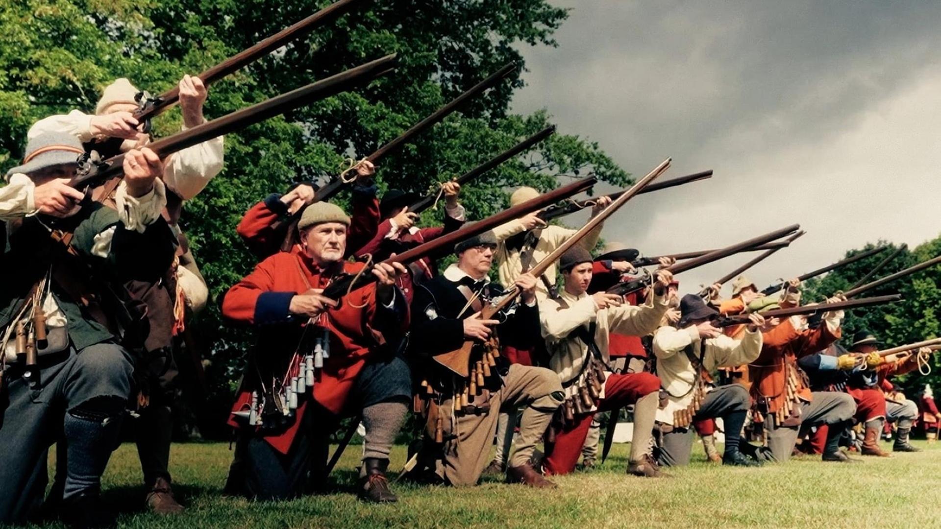 Auf einer Wiese kniet eine Gruppe von Männern in einer Reihe. Sie sind mit Musketen bewaffnet und halten die Gewehre im Anschlag.