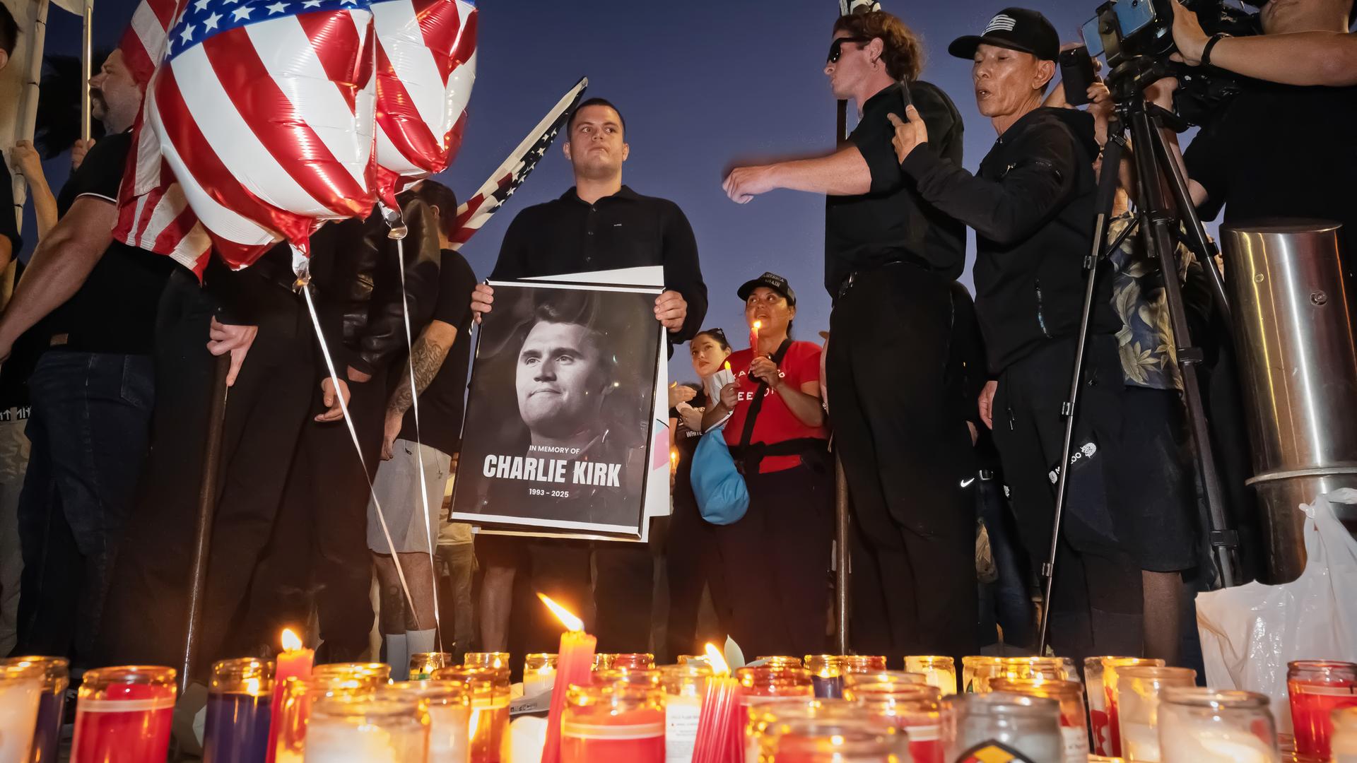 Männer in schwarzer Kleidung und mit US-Flaggen haben sich um Gedenkkerzen und das Schwarz-weiß-Foto eines Mannes versammelt.