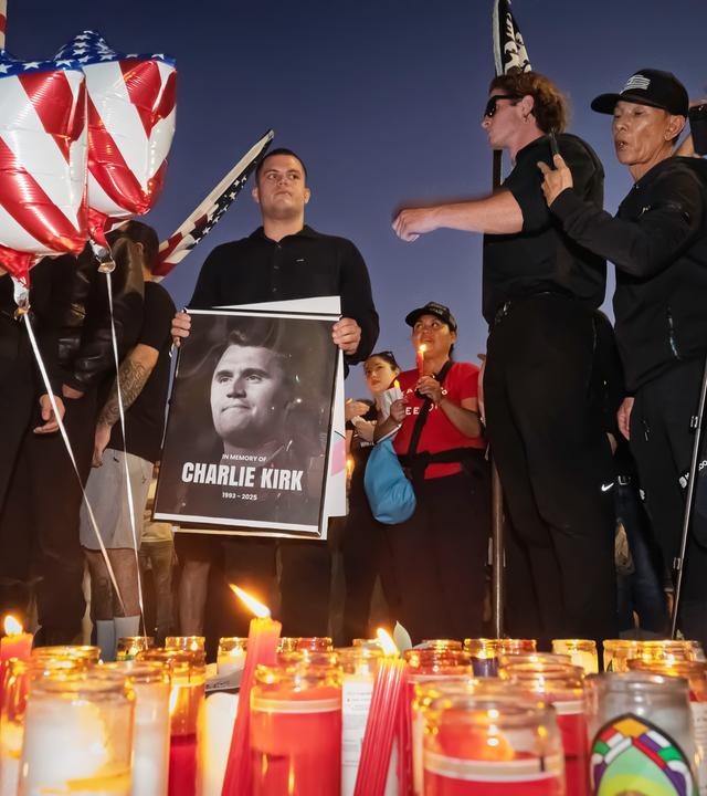 Männer in schwarzer Kleidung und mit US-Flaggen haben sich um Gedenkkerzen und das Schwarz-weiß-Foto eines Mannes versammelt.