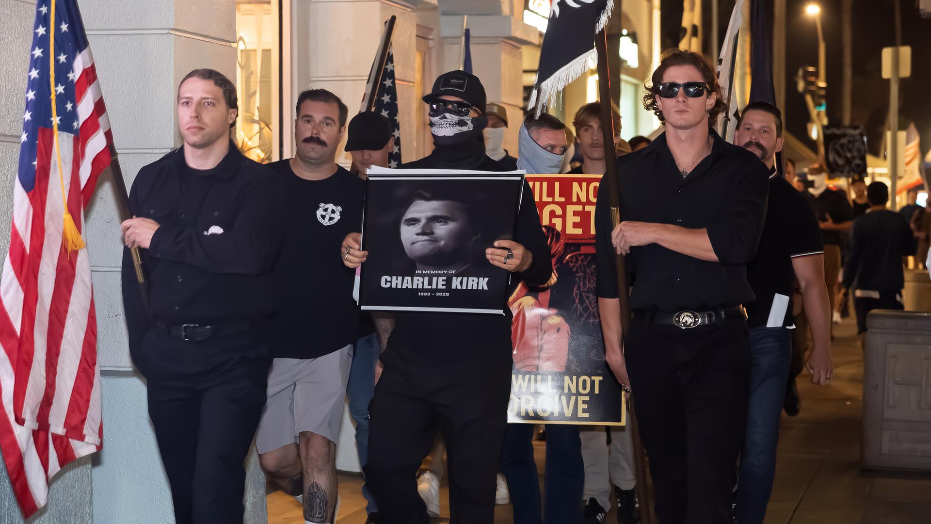 Männer in schwarzer Kleidung, teilweise vermummt, marschieren mit Fahnen und tragen das Porträtfoto eines Mannes vor sich her.