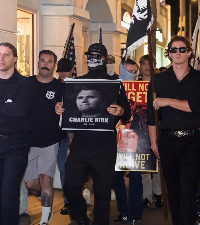 Männer in schwarzer Kleidung, teilweise vermummt, marschieren mit Fahnen und tragen das Porträtfoto eines Mannes vor sich her.