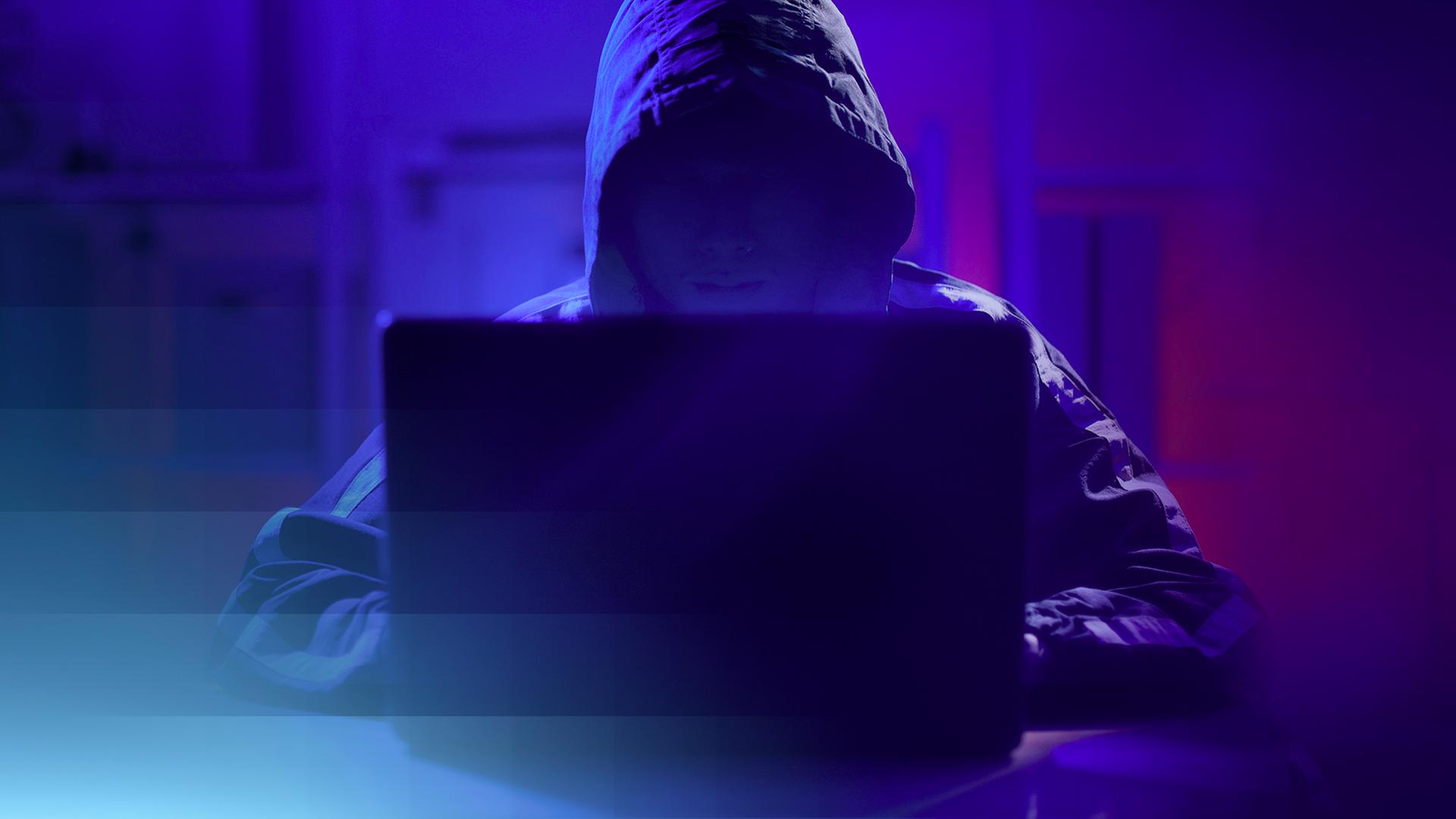 Beispielbild einer nicht erkennbaren Person mit Kapuze, die in blauem, gedimmten Licht vor einem Laptop sitzt.