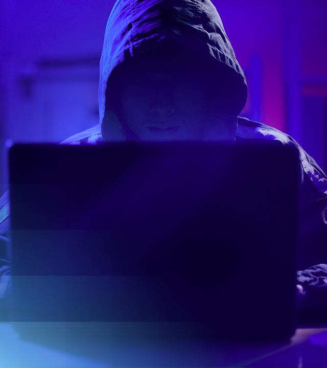 Beispielbild einer nicht erkennbaren Person mit Kapuze, die in blauem, gedimmten Licht vor einem Laptop sitzt.