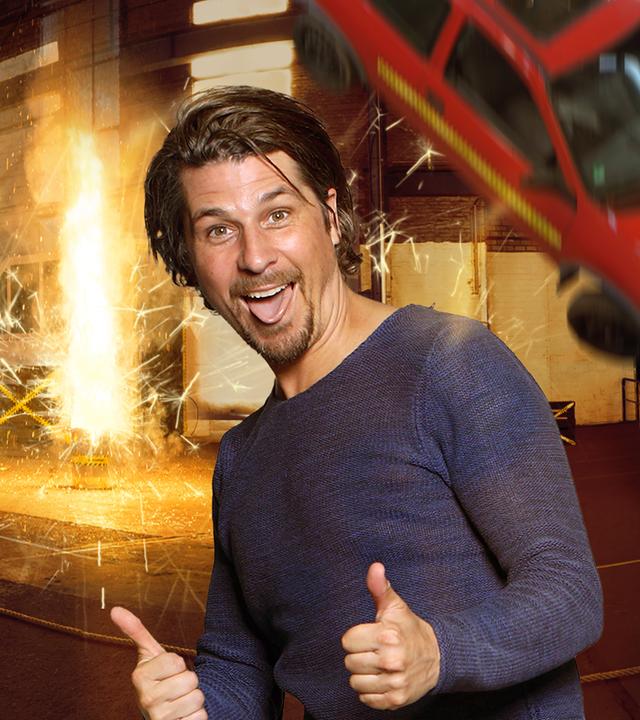 Checkpoint: Der Moderator der Sendung, Tommy Scheel, streckt die Zunge heraus und streckt beide Daumen nach oben. Ein grimmig schauender Junge rechts neben ihm hält eine Art Laubbläser und im Hintergrund stürzt ein Auto von der Decke.