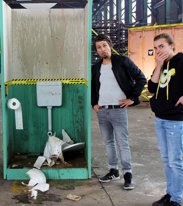 Tommy und das Team stehen um eine demolierte Kloschüssel herum.