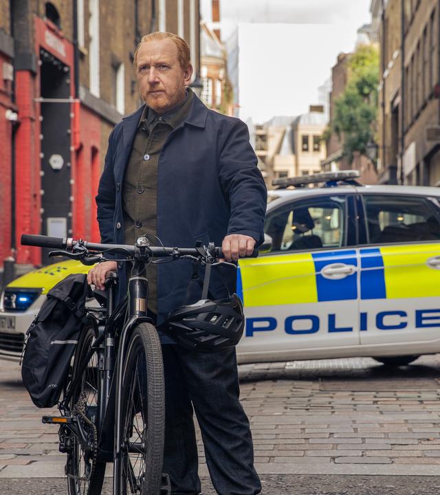 "The Chelsea Detective - Im blauen Zimmer": In einer kleinen Seitenstraße: DI Max Arnold (Adrian Scarborough) stellt sein Fahrrad auf der Straße ab. Sein Helm baumelt am Lenker. Im Hintergrund stehen zwei Polizeifahrzeuge.