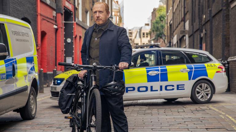 "The Chelsea Detective - Im blauen Zimmer": In einer kleinen Seitenstraße: DI Max Arnold (Adrian Scarborough) stellt sein Fahrrad auf der Straße ab. Sein Helm baumelt am Lenker. Im Hintergrund stehen zwei Polizeifahrzeuge.