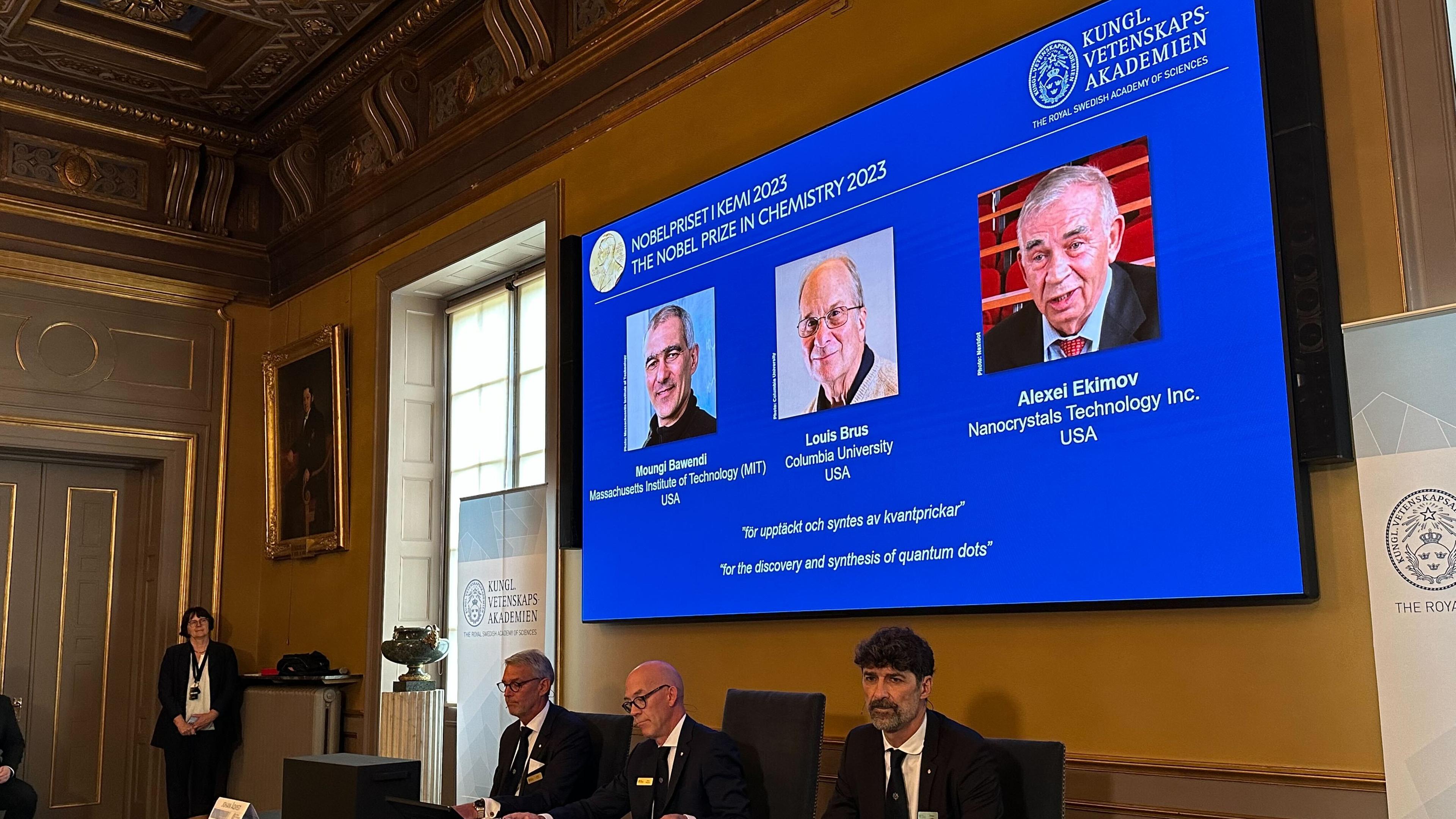 Drei Männer sitzten an einem Tisch, über ihnen eine Präsentation mit den Gewinnern des Chemie Nobelpreises.
