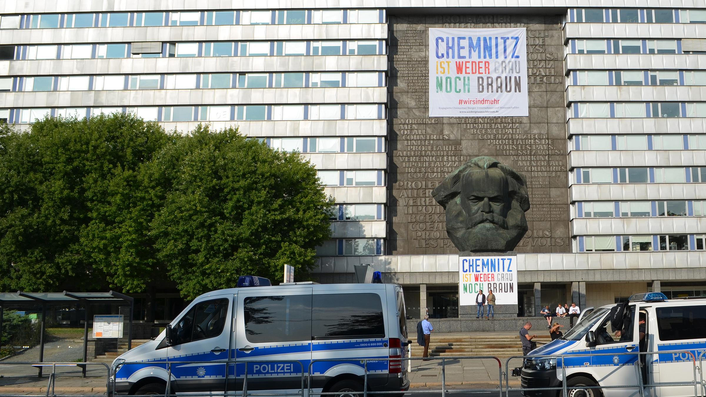 Abgesperrtes Karl-Marx-Monument in Chemnitz mit Plakat "Chemnitz ist weder Grau noch Braun" und Polizeiwagen im Vordergrund.