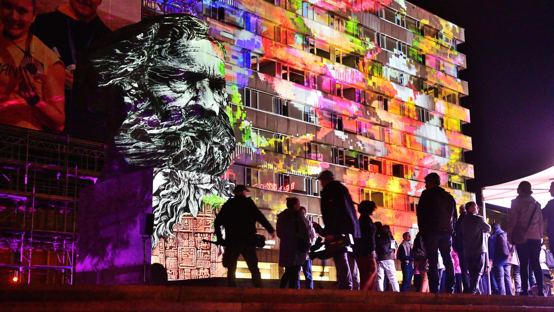 Der große Karl-Marx-Kopf vor einem modernen Gebäude wird nachts mit bunten Lichtern angeleuchtet. Davor sieht man die dunklen Silhouetten von Menschen.