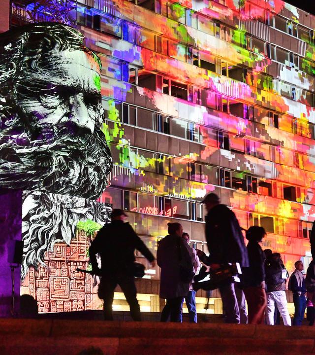 Der große Karl-Marx-Kopf vor einem modernen Gebäude wird nachts mit bunten Lichtern angeleuchtet. Davor sieht man die dunklen Silhouetten von Menschen.