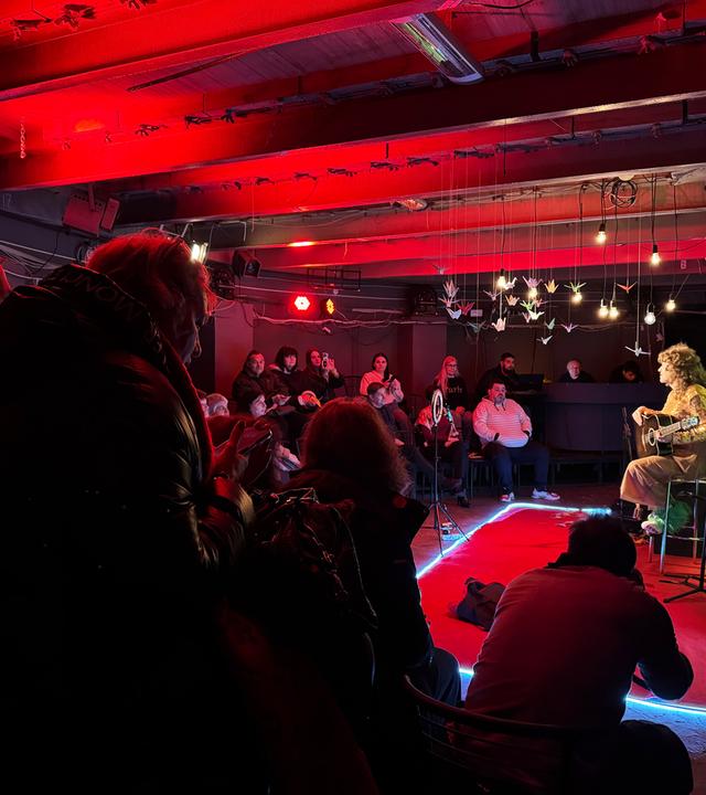 In einem rot ausgeleuchteten Kellerraum spielt eine Person auf der Bühne Gitarre, während rundrum Menschen im Halbdunklen sitzen und zuhören.