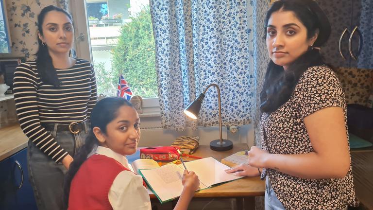Szene aus "Western Lane" von Chetna Maroo. Ein junges Mädchen sitzt an einem Schreibtisch mit einem aufgeschlagenen Heft und einem Stift in der Hand. Rechts und links von ihr stehen zwei jugendliche Frauen mit einem ernsten Gesichtsausdruck.