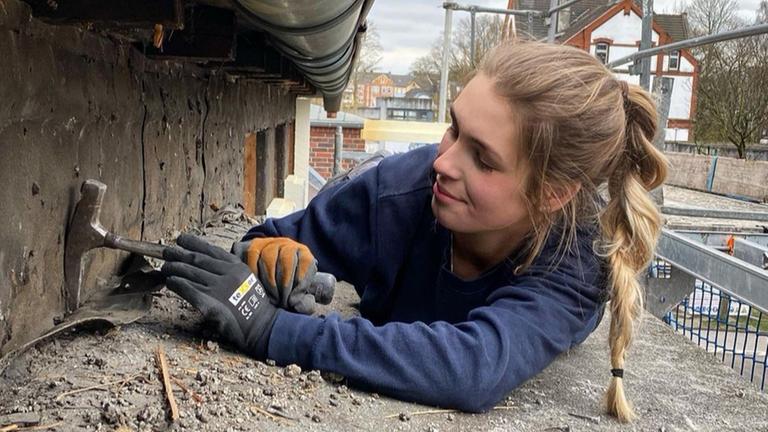 Eine junge Frau arbeitet auf dem Bauch liegend unter einer Dachtraufe.