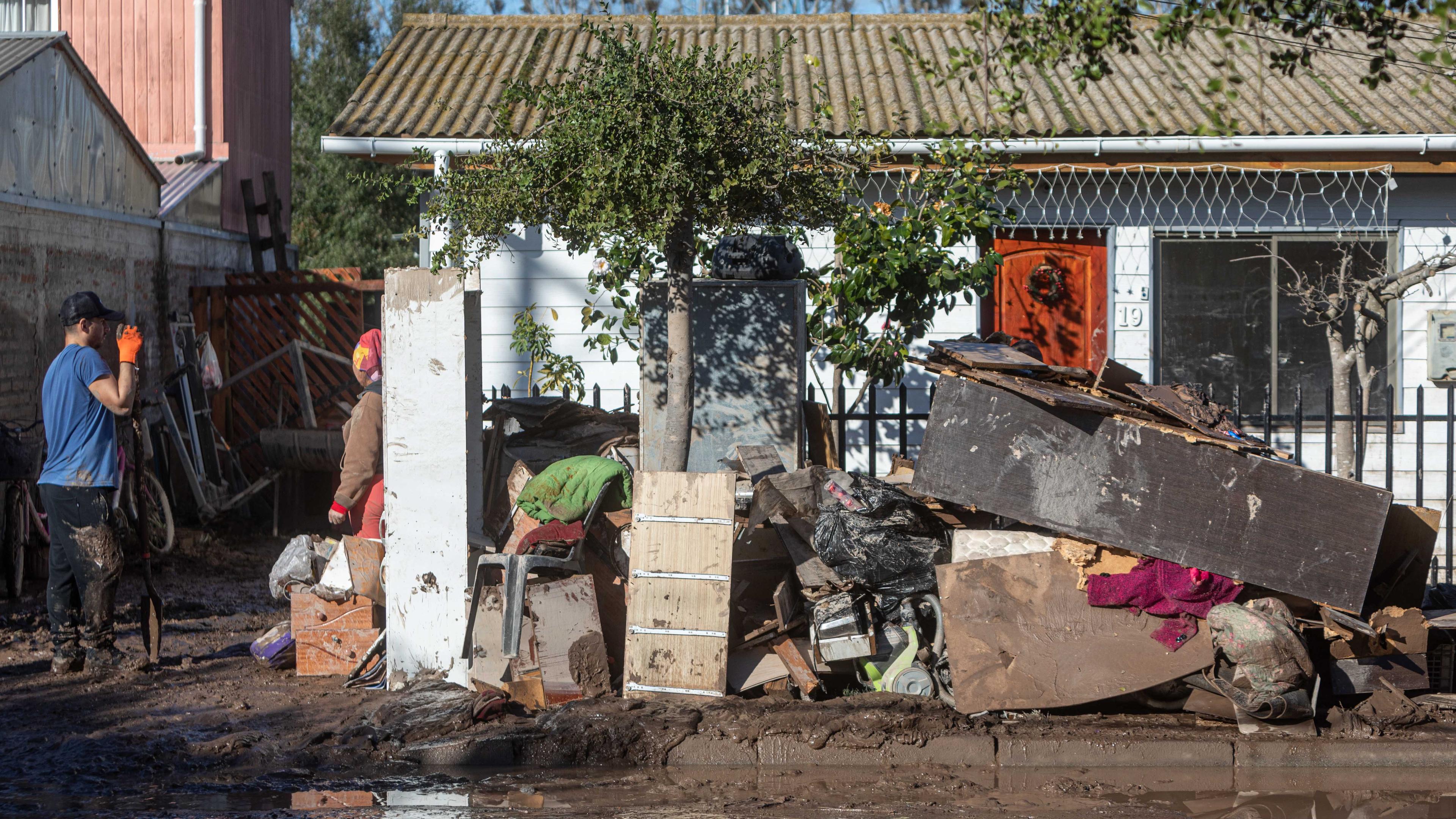 Trümmer auf der Straße nach einer Überschwemmung in Chile. Links im Bild Personen bei Aufräumarbeiten. Im Hintergrund ein Haus.