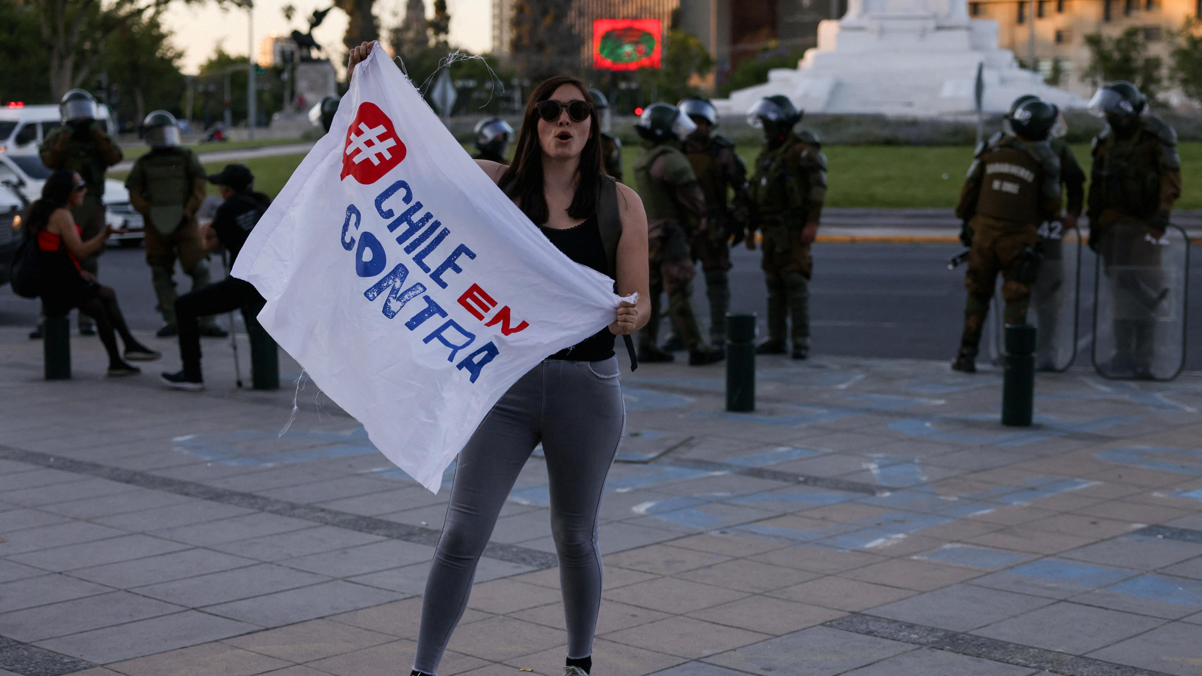 Eine Demosntrantin hält ein Plakat mit der Aufschrift "Chile en contra", im Hintergrund stehen Polizeibeamte