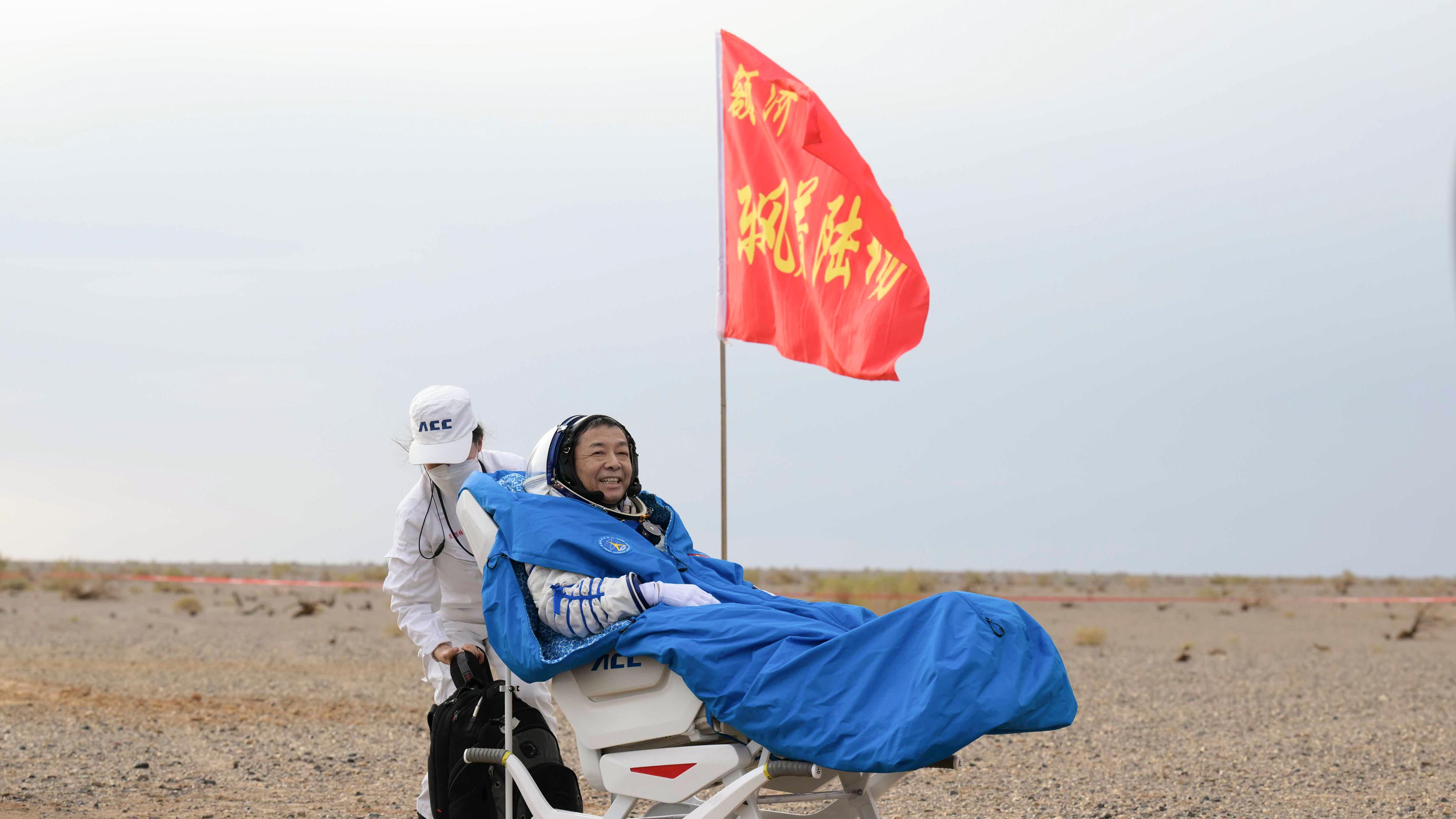 Der chinesische Astronaut Deng Qingming sitzt lachend auf einer Trage in der Wüste, neben ihm weht eine rote Flagge. 