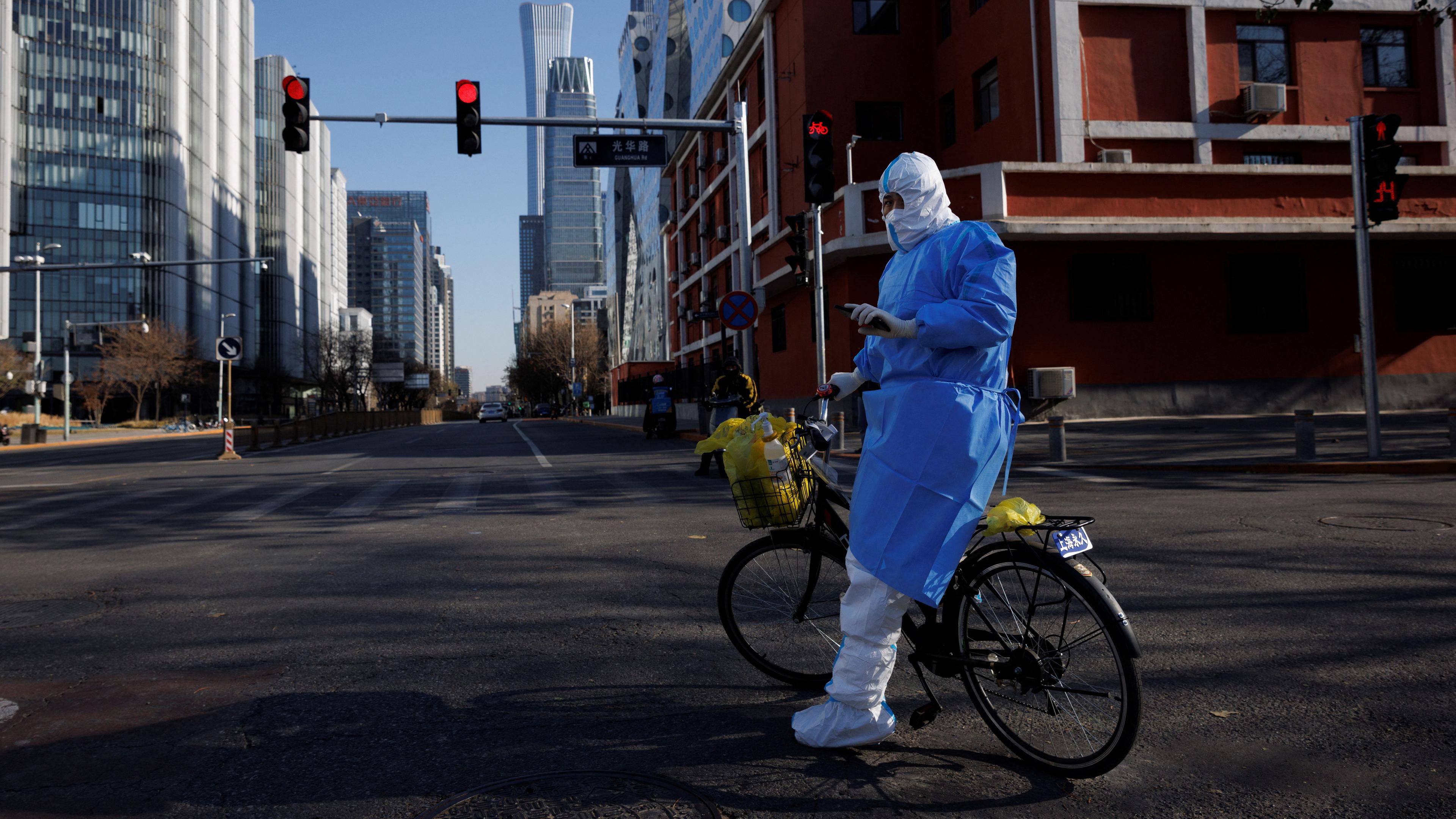Eine Person in einem blauen Schutzanzug steht an ein Fahrrad gelehnt auf einer leeren Straße in Peking.