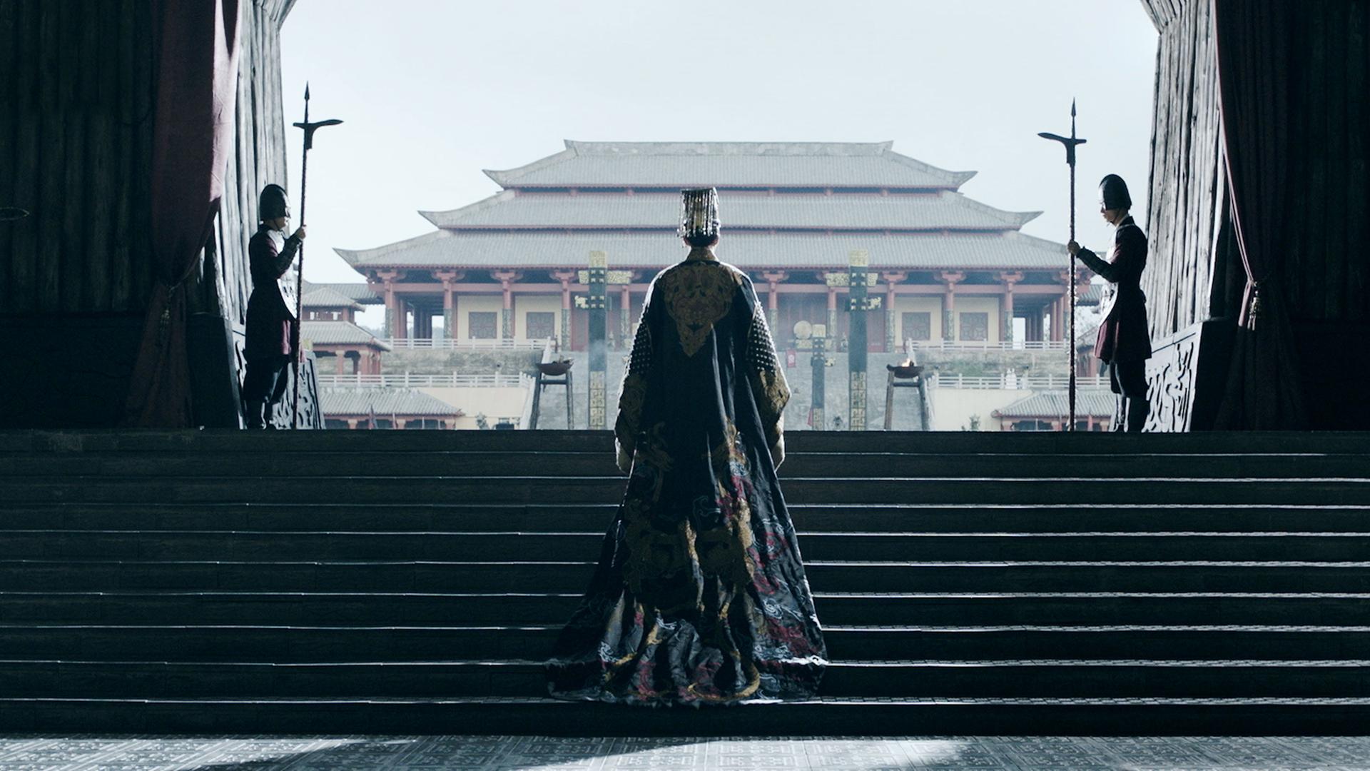  Spielszene. Ein Mann in langer Seidenrobe in Rückenansicht steigt die ersten Stufen einer Treppe hinauf, die aus einem im Halbdunkel liegenden Raum auf eine Terrasse führt. Der Blick öffnet sich auf eine Pagode im Hintergrund, darüber der hellblaue Himmel. Links und rechts begrenzen die Seitenwände des Portals den Ausblick. Zwei Soldaten stehen mit aufgestellten Lanzen links und rechts am unteren Ende der Treppe, zwei weitere auf dem oberen Absatz der Treppe.
