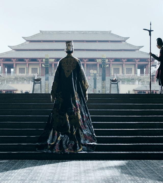  Spielszene. Ein Mann in langer Seidenrobe in Rückenansicht steigt die ersten Stufen einer Treppe hinauf, die aus einem im Halbdunkel liegenden Raum auf eine Terrasse führt. Der Blick öffnet sich auf eine Pagode im Hintergrund, darüber der hellblaue Himmel. Links und rechts begrenzen die Seitenwände des Portals den Ausblick. Zwei Soldaten stehen mit aufgestellten Lanzen links und rechts am unteren Ende der Treppe, zwei weitere auf dem oberen Absatz der Treppe.