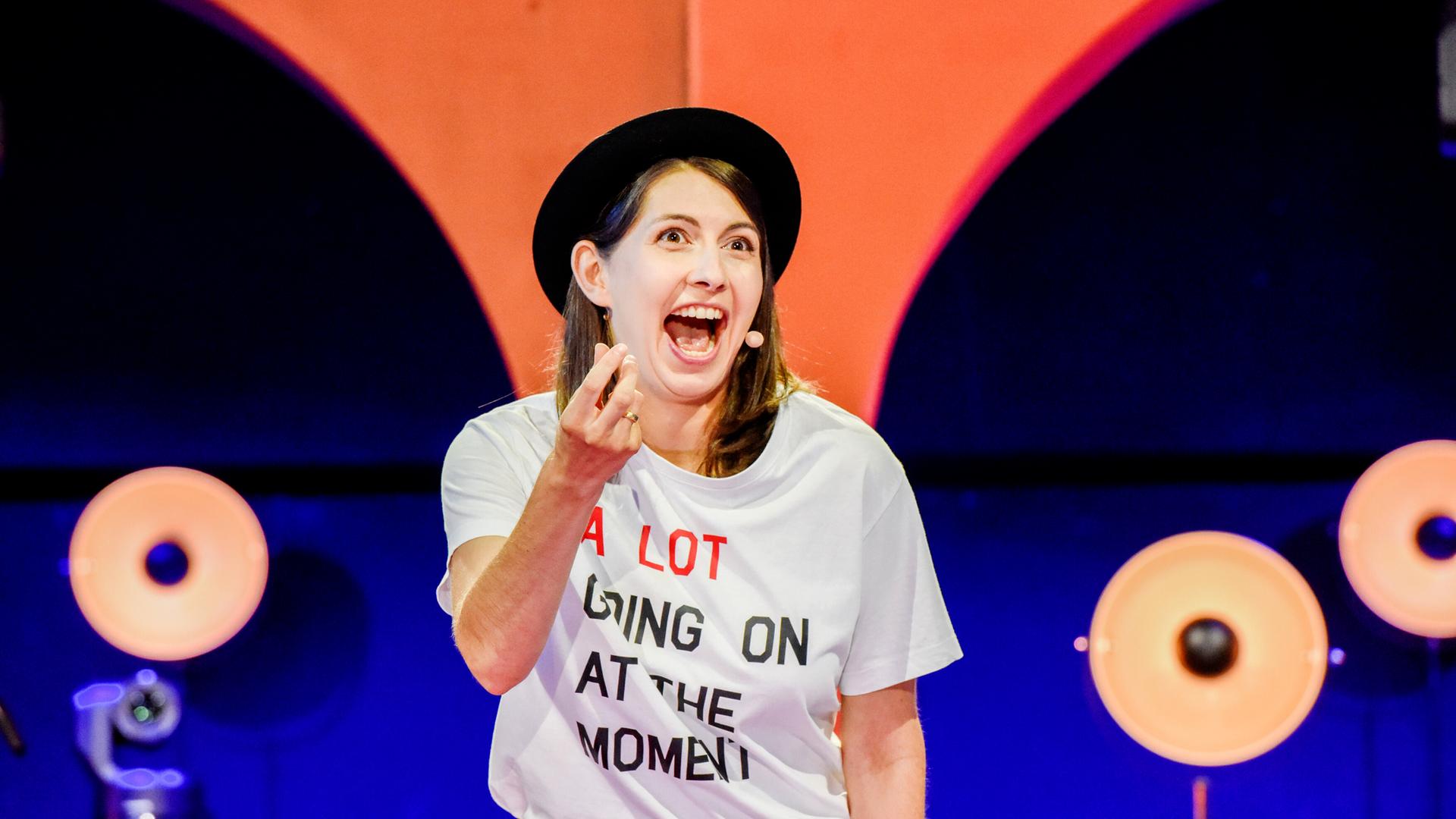 Eine Frau mit schwarzem Hut und weissem beschrifteten T-Shirt vor einer Bühnendeko in blau/orange