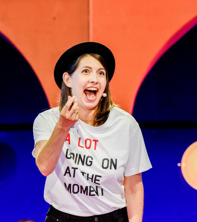 Eine Frau mit schwarzem Hut und weissem beschrifteten T-Shirt vor einer Bühnendeko in blau/orange