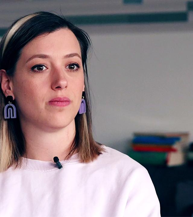 Eine traurig blickende Frau in einem rosafarbenen Pulli und mit lila Ohrringen.