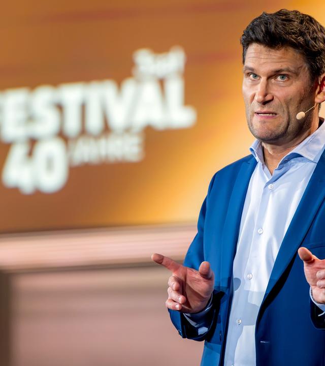 Christian Ehring steht auf der rechten Seite des Bildes. Er hebt beide Zeigefinger. Der Hintergrund ist gelb und braun beleuchtet. Auf der linken Bildseite ist im Hintergrund das 3satFestival Logo abgebildet. Christian Ehring trägt einen dunkelblauen Anzug.