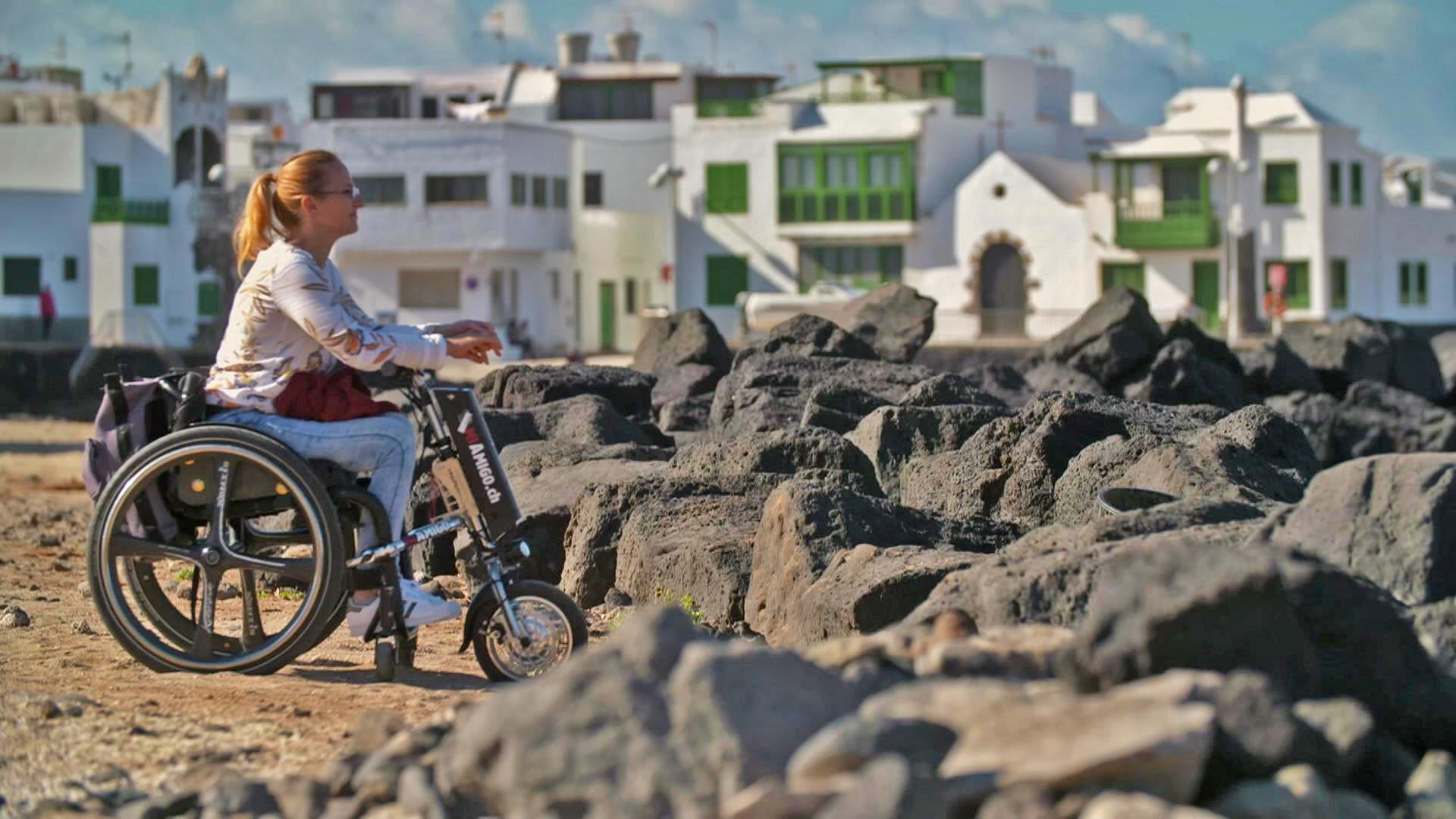 Christiane Göldner sitzt im Rollstuhl vor schwarzen Felsen, im Hintergrund Häuser.