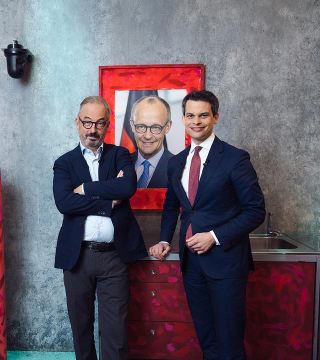 Das Bild zeigt den CDU-Politiker Christoph Ploß und Jan Fleischhauer in einer stilisierten Raumkulisse mit grauer Betonwand. Beide stehen nebeneinander vor einer kleinen Küchenzeile und einem auffällig rot bemalten Kühlschrank. Hinter ihnen hängt ein großformatiges Porträt von Friedrich Merz in einem rot gestalteten Rahmen.
