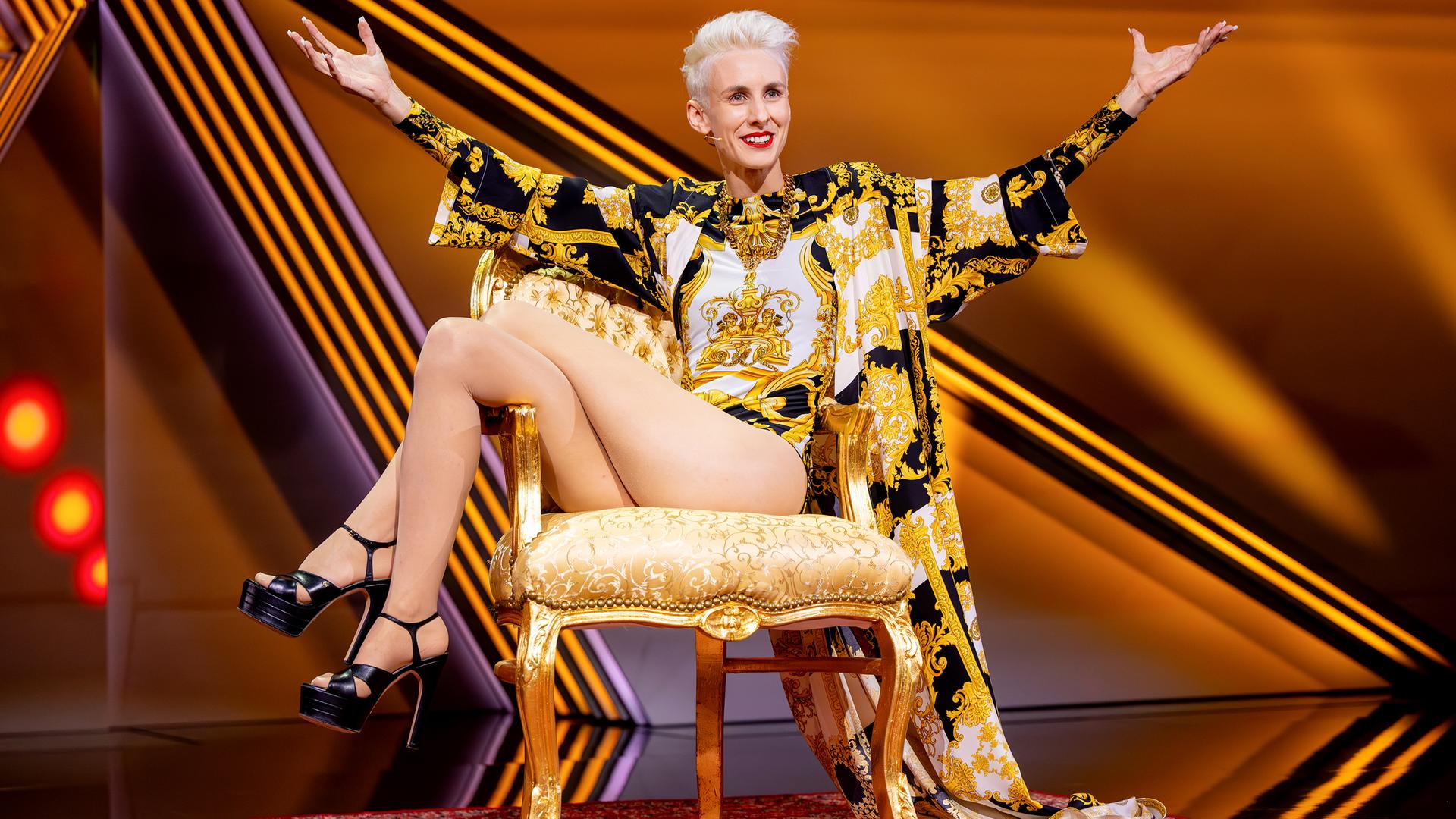 Kabarettistin itzt in der Mitte des Bildes auf einem goldenen Stuhl. Sie breitet die Hände zur Seite aus und streckt die Beine zur linken Bildseite. Der Hintergrund ist orange beleuchtet.