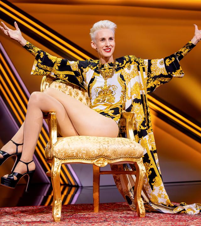 Kabarettistin itzt in der Mitte des Bildes auf einem goldenen Stuhl. Sie breitet die Hände zur Seite aus und streckt die Beine zur linken Bildseite. Der Hintergrund ist orange beleuchtet.
