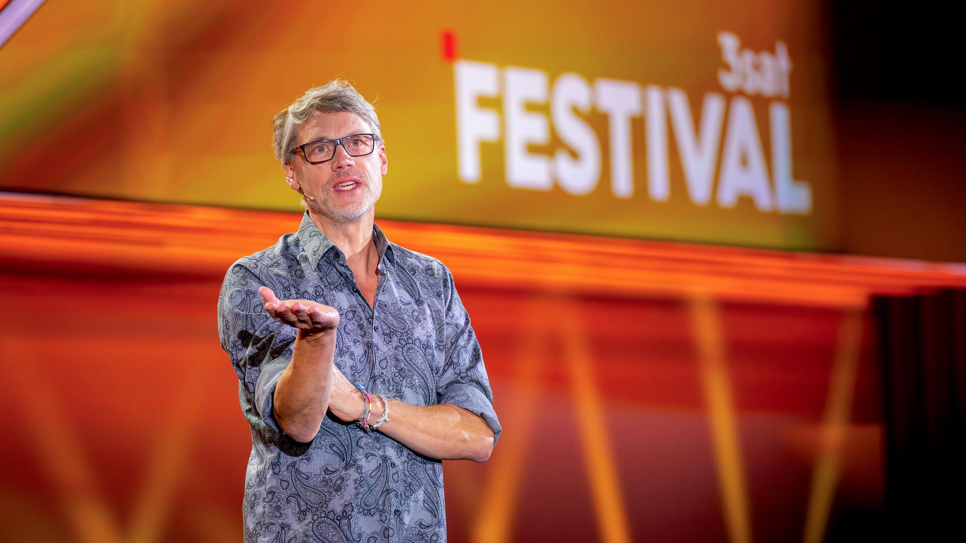 Kabarettist steht auf der linken Seite des Bildes. Er trägt eine Brille. Seine rechte Hand streckt er nach vorne. Er trägt ein gemustertes Hemd. Der Hintergrund ist orange und rot beleuchtet.