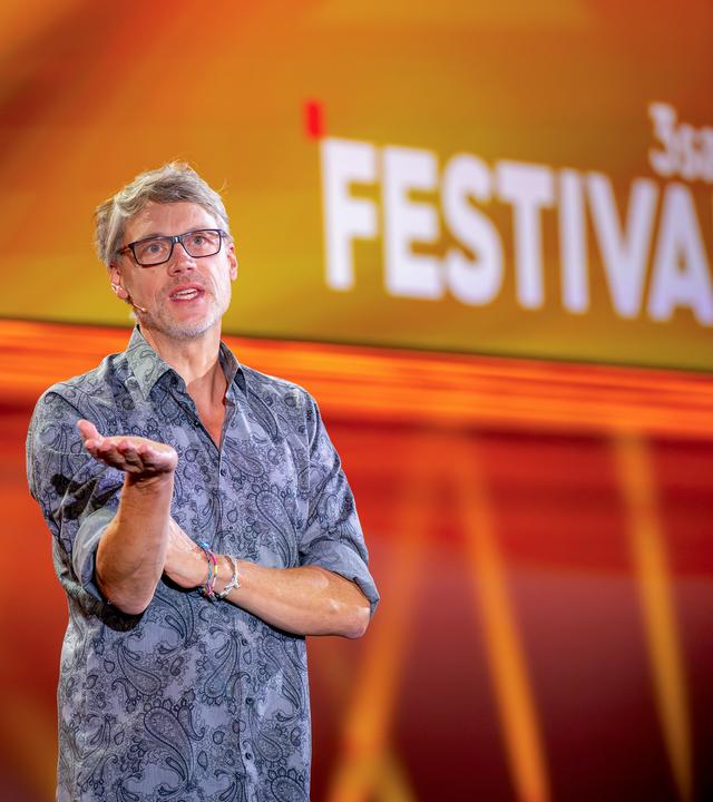 Kabarettist steht auf der linken Seite des Bildes. Er trägt eine Brille. Seine rechte Hand streckt er nach vorne. Er trägt ein gemustertes Hemd. Der Hintergrund ist orange und rot beleuchtet.